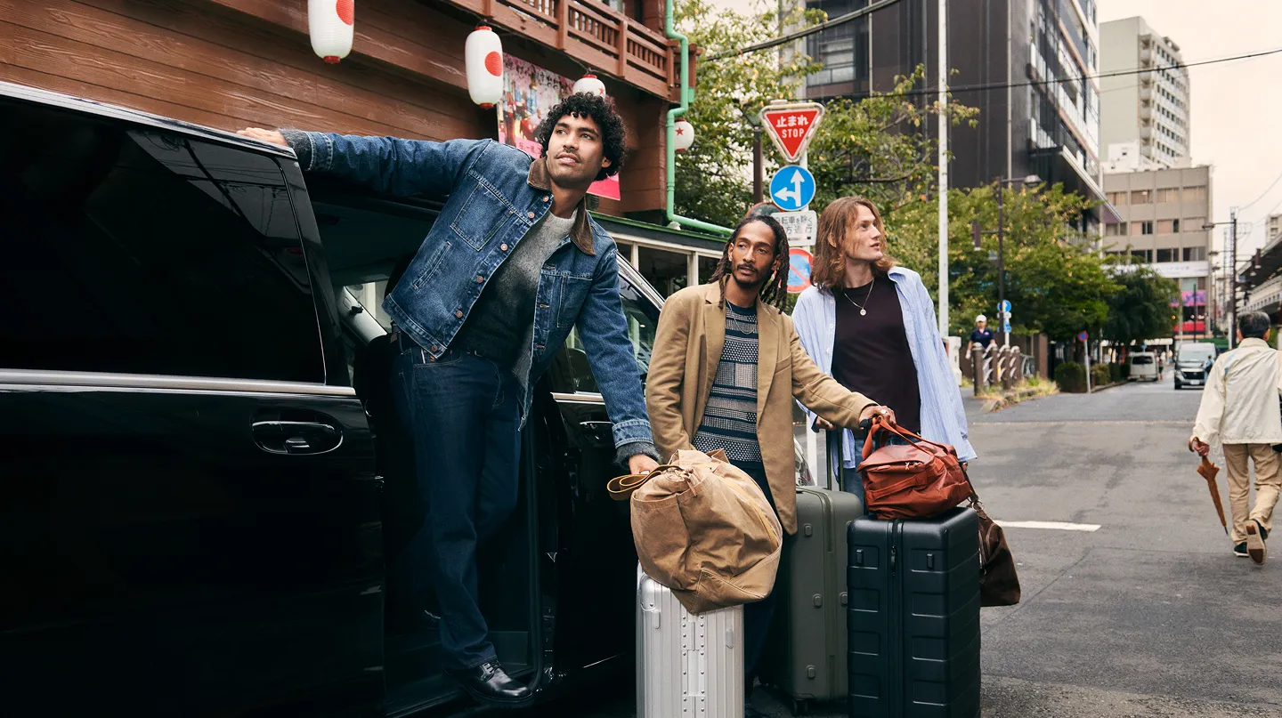 Three men standing beside a car, with luggage placed on the ground next to them, ready for travel.